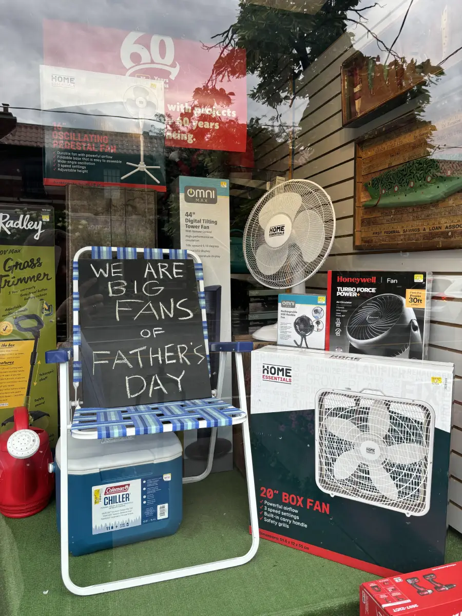 Store window display features various fans, a cooler, and a lawn chair with a sign saying “We are BIG FANS of Father’s Day.” The setup promotes gifts for Father’s Day, with reflections of trees in the glass.