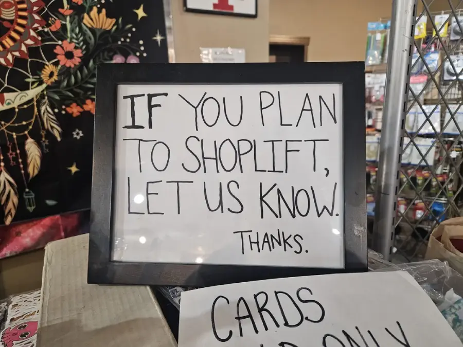 A framed sign reads, “If you plan to shoplift, let us know. Thanks.” It is displayed on a counter with a tapestry and greeting cards visible in the background.
