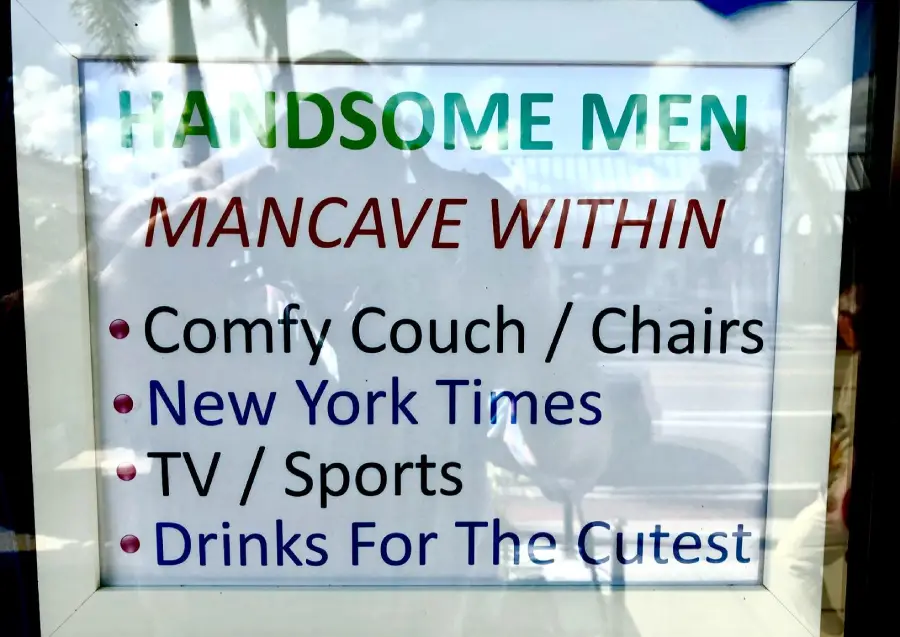 A sign reads: “HANDSOME MEN MANCAVE WITHIN. Comfy Couch / Chairs, New York Times, TV / Sports, Drinks For The Cutest,” with colorful and bold text styles.