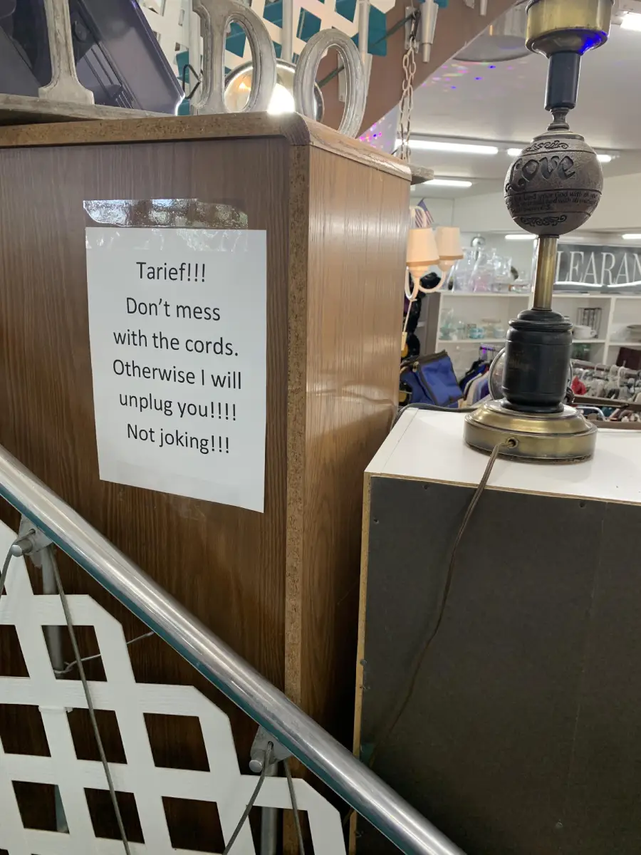 A sign on a wooden cabinet warns, “Tarief!!! Don’t mess with the cords. Otherwise I will unplug you!!! Not joking!!!” Near it is a lamp with a cord and some store items in the background.