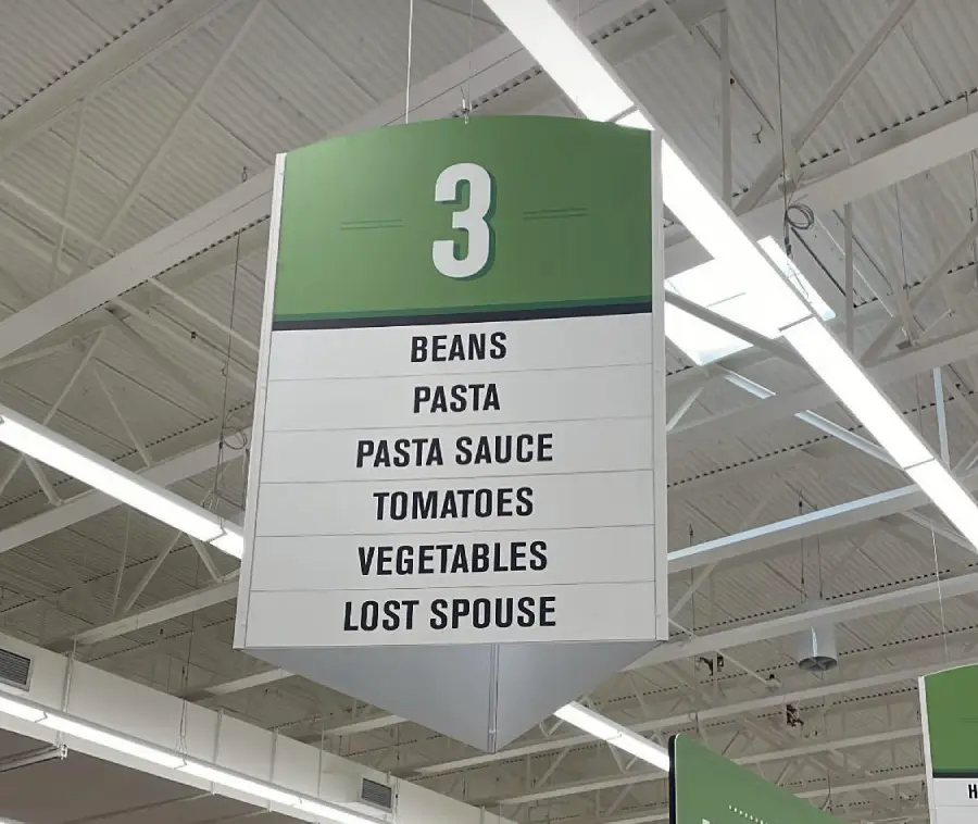 A grocery store aisle sign labeled “3” lists beans, pasta, pasta sauce, tomatoes, vegetables, and humorously, “lost spouse” under the items found in that aisle.