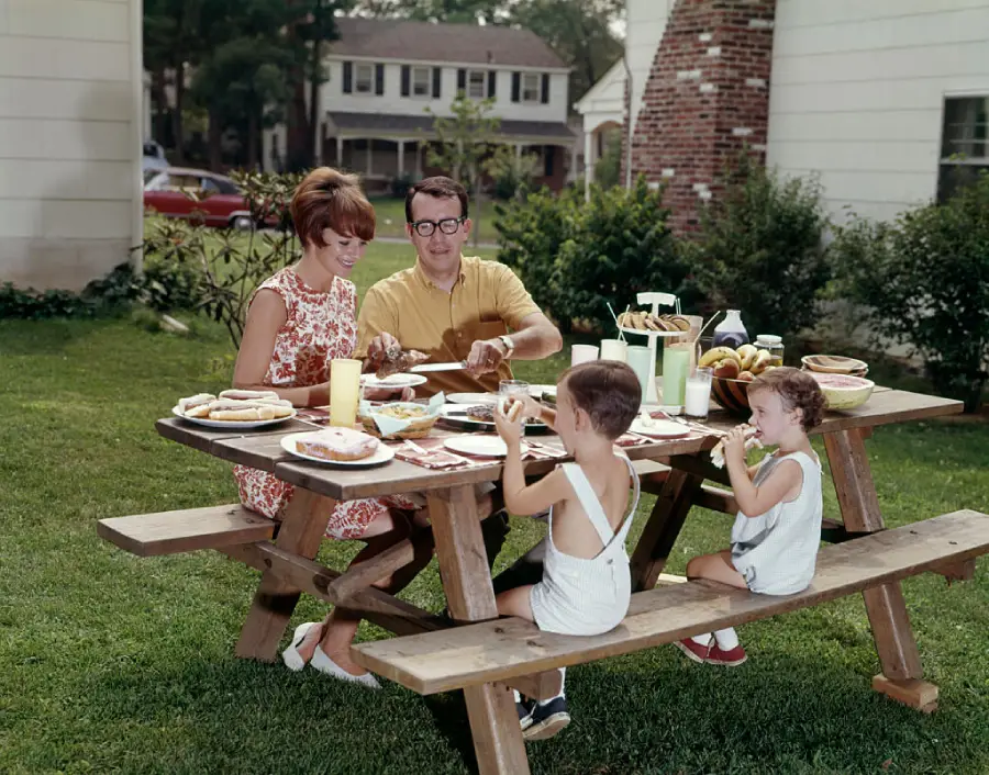 A family of four sits at a wooden picnic table in a backyard, enjoying a meal together on a sunny day. The parents smile as the father serves food, while two young children eat sandwiches.
