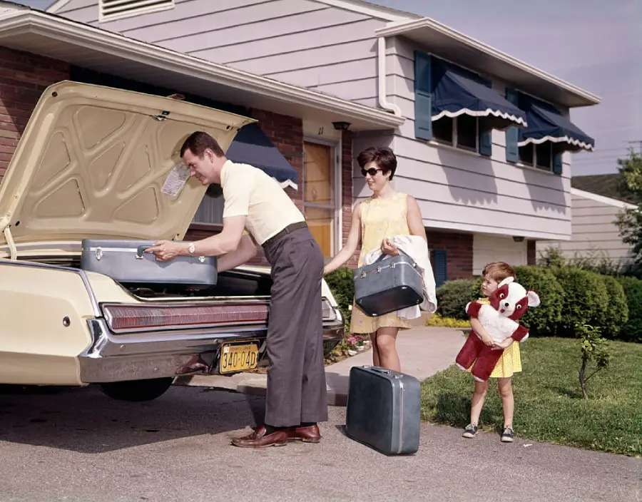 A man and woman load suitcases into a car trunk while a young girl holding a teddy bear watches, standing outside a suburban house on a sunny day.