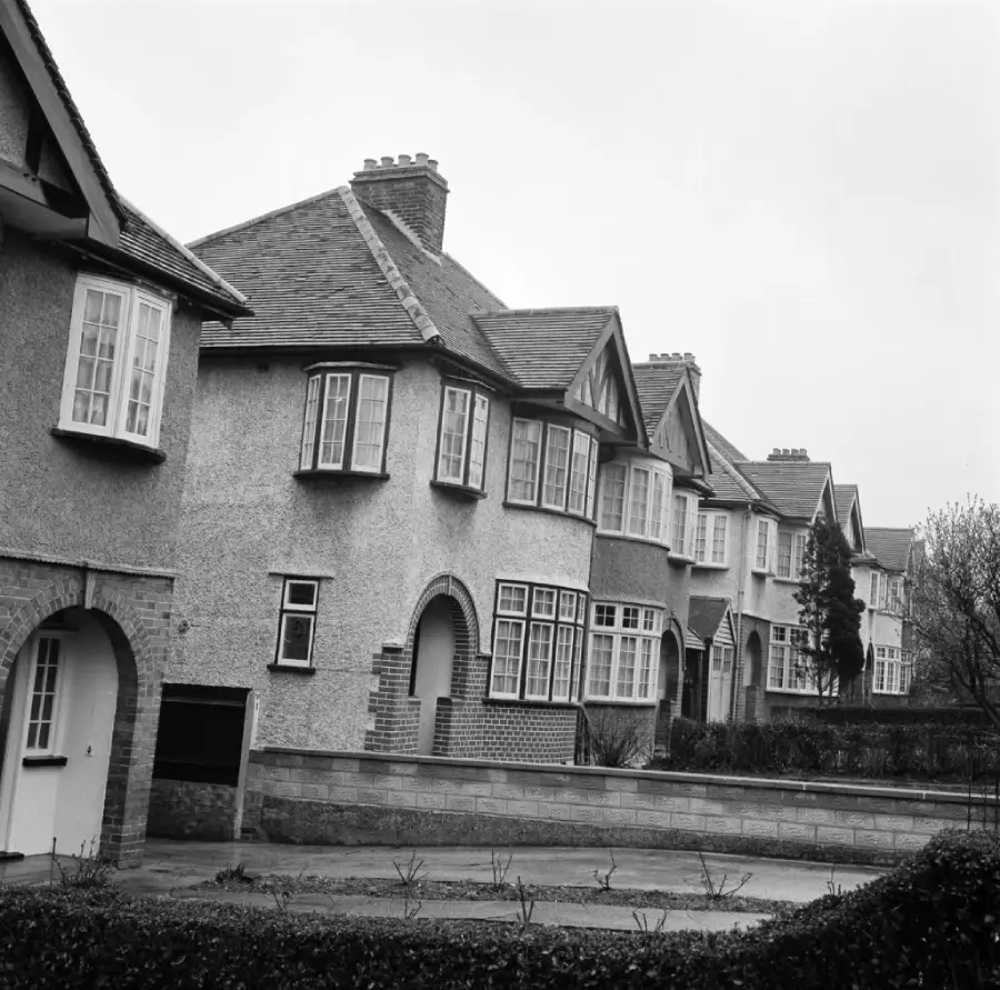 A row of semi-detached suburban houses with bay windows, pitched roofs, and chimneys. The scene is in black and white, suggesting an older or vintage photograph. Bushes and a stone wall line the front of the homes.