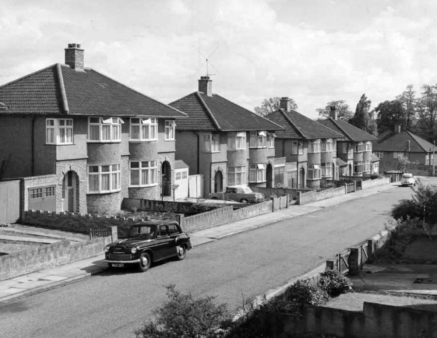 A black-and-white photo of a quiet suburban street lined with semi-detached houses and a classic car parked along the curb, evoking a mid-20th century atmosphere.