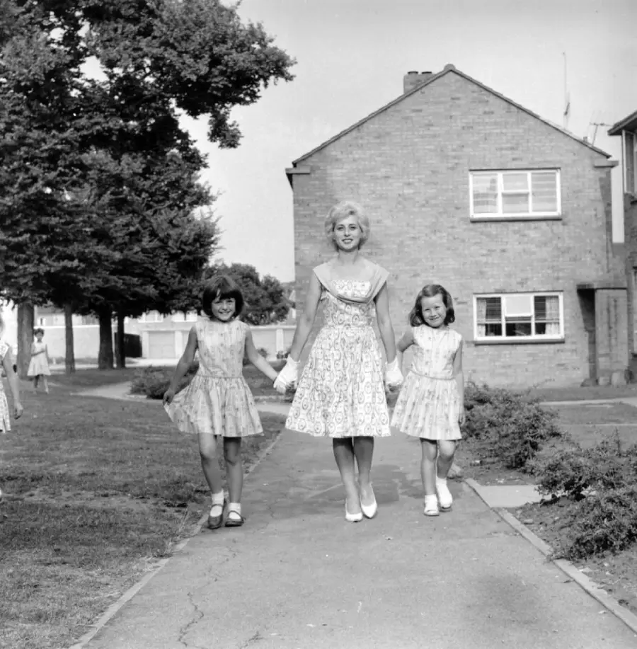 A woman in a patterned dress smiles while walking outside, holding hands with two young girls in matching dresses and shoes, on a paved path near a brick house and trees.