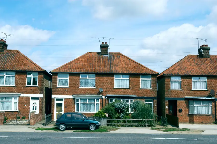 A row of three brick houses with red-tiled roofs, white windows, and small front gardens. A blue car is parked in front on the street under a blue sky with scattered clouds.