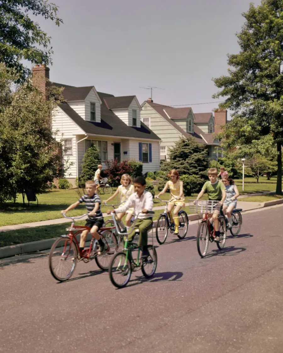 Five children ride bicycles along a suburban street lined with houses and trees on a sunny day. The kids appear to be enjoying themselves as they bike together in a group.