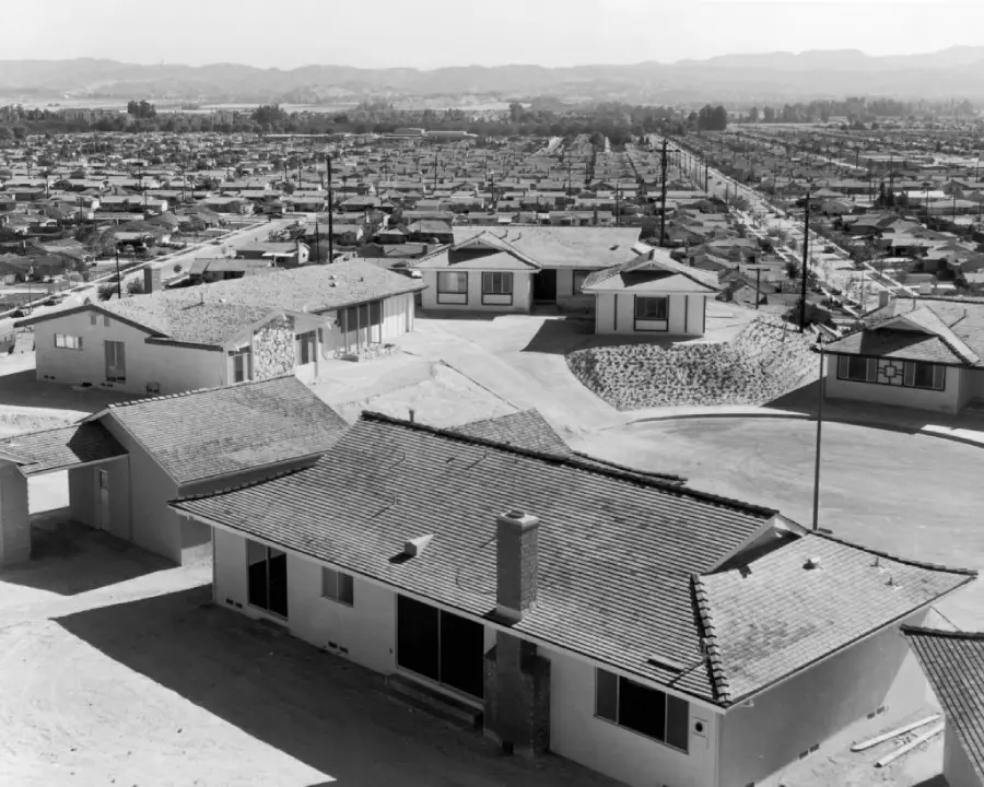 Black-and-white photo of a suburban neighborhood with rows of identical single-story houses, arranged in a grid pattern, seen from an elevated viewpoint.
