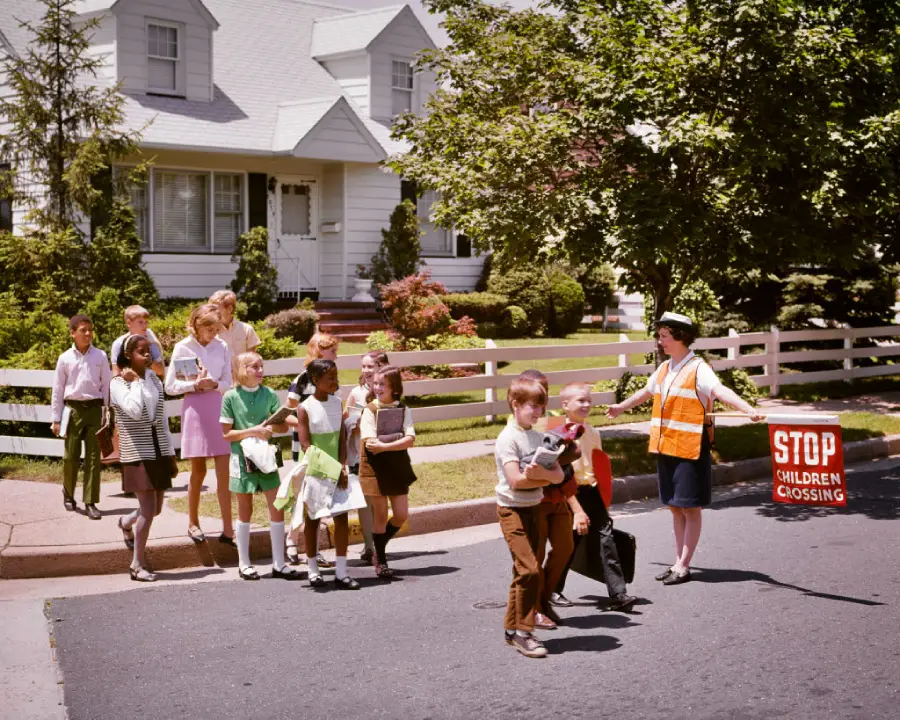 A crossing guard holds a “STOP Children Crossing” sign, helping a group of schoolchildren cross a suburban street in front of white houses and a picket fence on a sunny day.