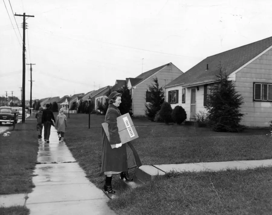 A woman in a coat and hat carries a box labeled “RECORD LOAN” on a suburban sidewalk. Two people with umbrellas walk in the background, and houses line the street on a rainy day.