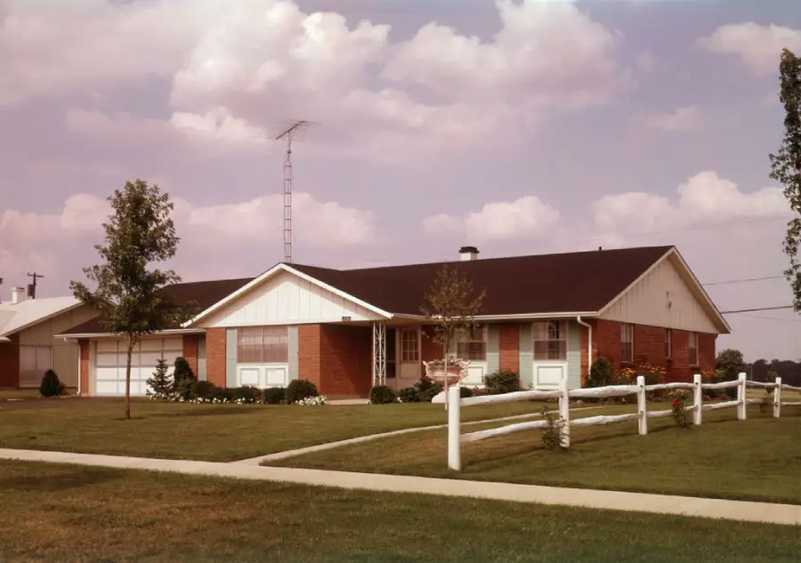 A single-story brick house with a dark roof, white trim, and a large front yard, surrounded by a white wooden fence. A metal TV antenna is mounted on the roof. The sky is partly cloudy.