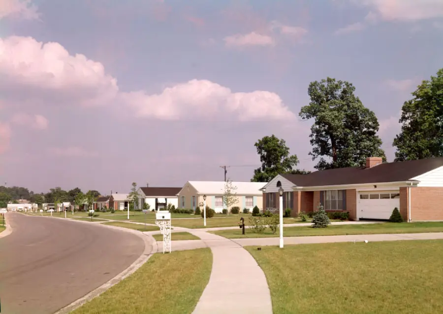 A suburban street with single-story houses, green lawns, and trees under a partly cloudy sky. The sidewalk curves alongside the road, and mailboxes are visible near driveways.