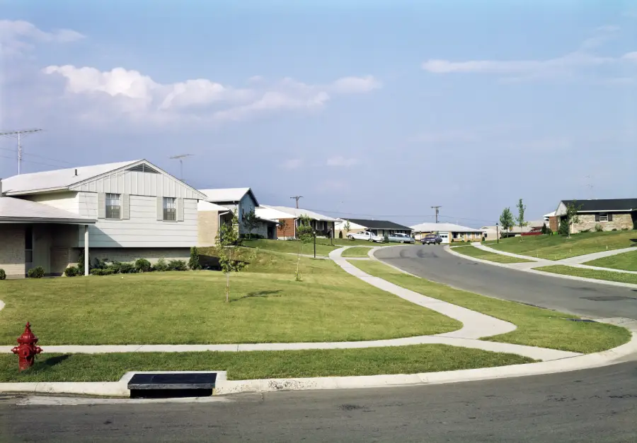 A suburban street with single-story houses, neatly trimmed lawns, a red fire hydrant, curved sidewalks, and a blue sky with scattered clouds.