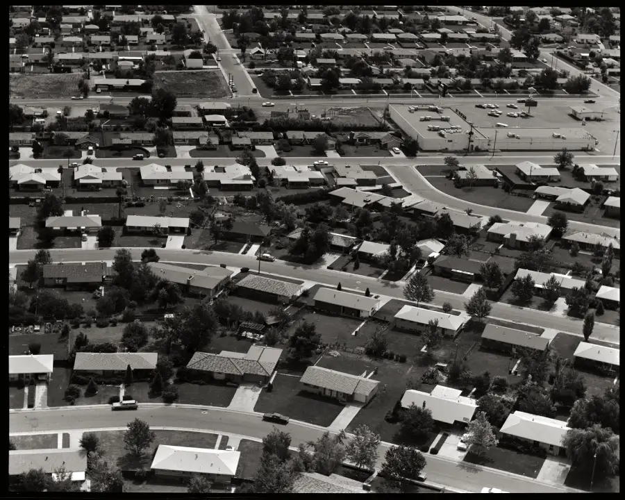 Aerial view of a suburban neighborhood with rows of single-story houses, green lawns, trees, curved streets, and a parking lot in the background.