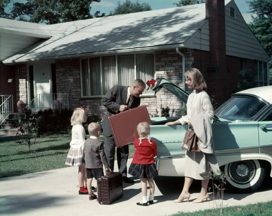 A man and woman load luggage into a vintage car’s trunk while three young children stand nearby in front of a suburban house.
