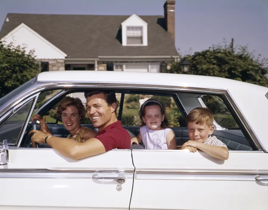 A family of four sits in a white car parked in front of a house. The father smiles at the wheel, the mother sits beside him, and two young children look out the open back window, all appearing happy.