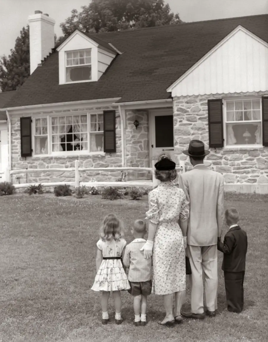 A family of four, dressed in 1950s clothing, stands on a lawn facing a stone house with shuttered windows and a chimney, viewing it from behind.