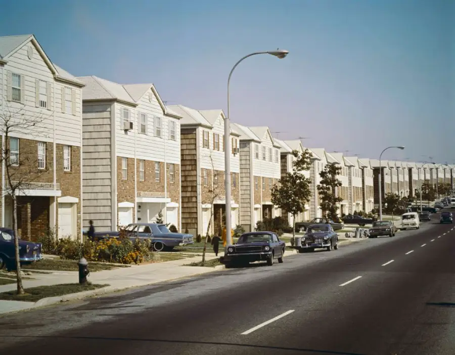 A row of identical three-story townhouses lines a wide suburban street, with vintage 1960s cars parked along the curb and a few small trees dotting the sidewalk under a clear, blue sky.