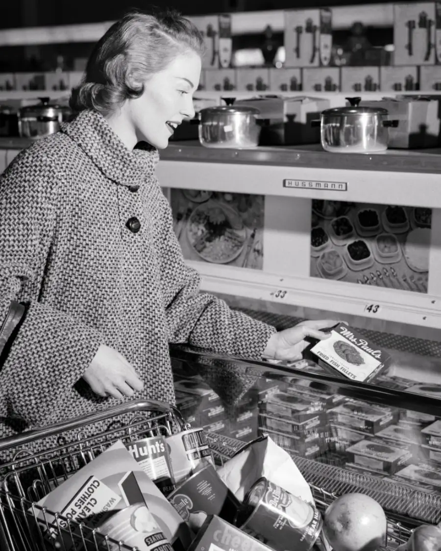 A woman in a coat smiles while shopping at a grocery store, holding a frozen meal above a freezer. Her cart is filled with groceries, including canned goods and produce.