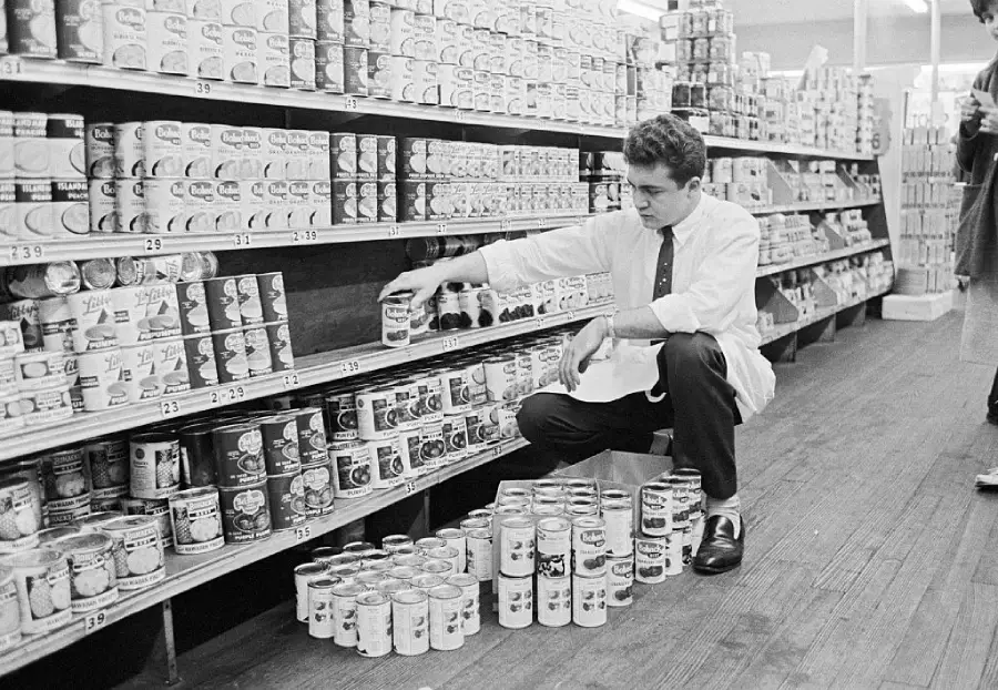 A man in a white shirt squats in a grocery store aisle, stocking shelves with canned goods. Stacks of cans are on the wooden floor and shelves are filled with various canned products.