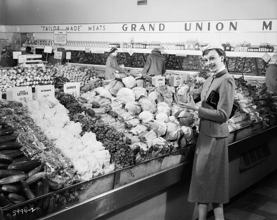 A woman in a suit and hat stands by a produce display in a grocery store, holding a cabbage. Other shoppers are visible in the background near signs for lettuce, carrots, and turnips. The sign above reads “GRAND UNION.”