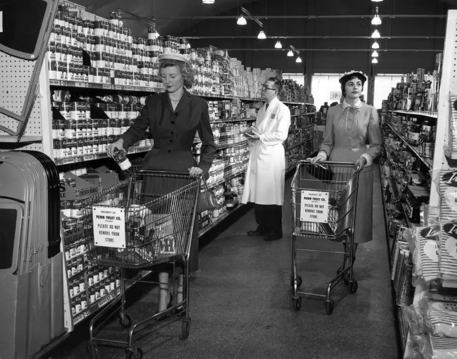 Two women with shopping carts browse grocery store shelves, while a man in a white coat stands in the aisle. The store appears vintage, and the shelves are stocked with canned goods. Bright lights hang from the ceiling.
