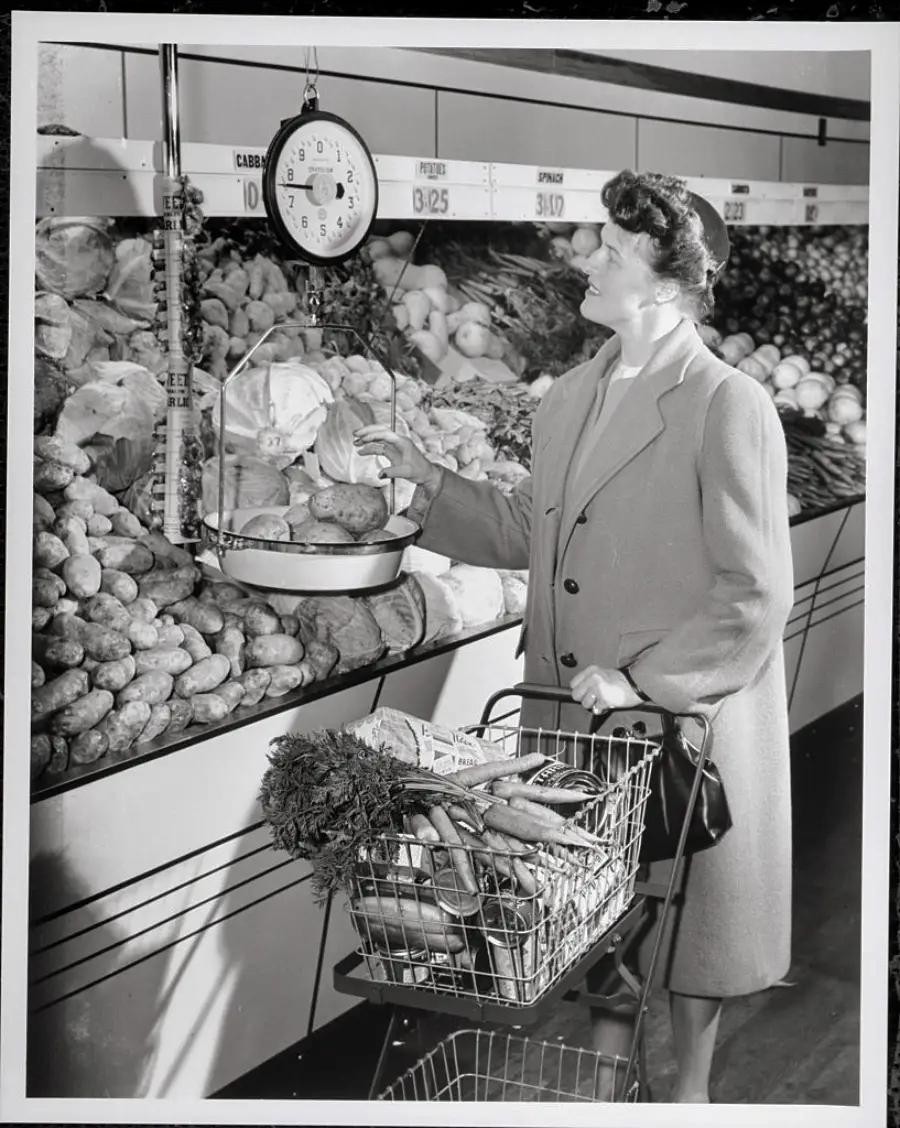 A woman in a coat weighs vegetables on a hanging scale in a grocery store produce section, with a basket of carrots and other items in her shopping cart.