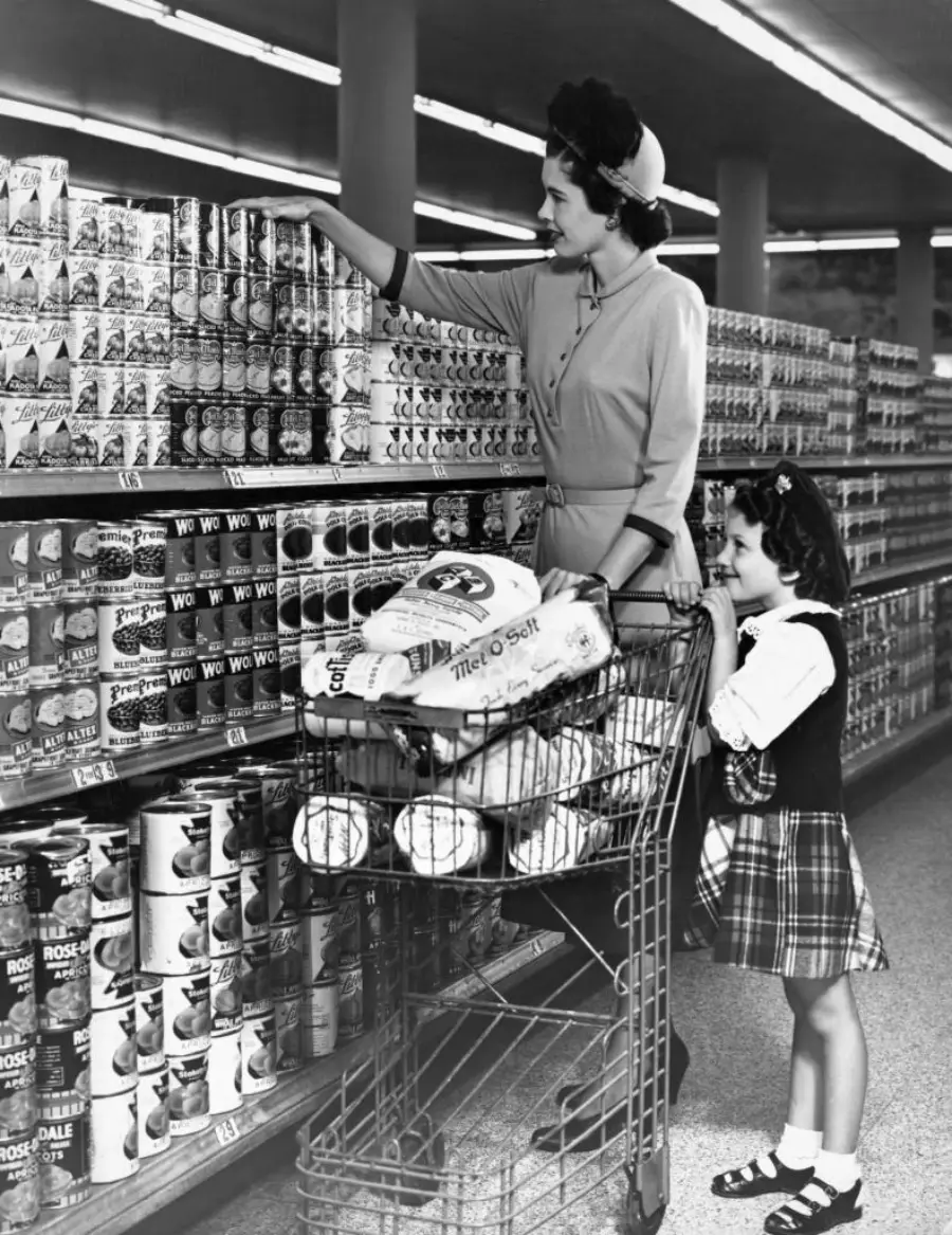 A woman and a young girl shop in a grocery store, picking canned goods from shelves. Their cart is filled with groceries, including large bags and loaves of bread.