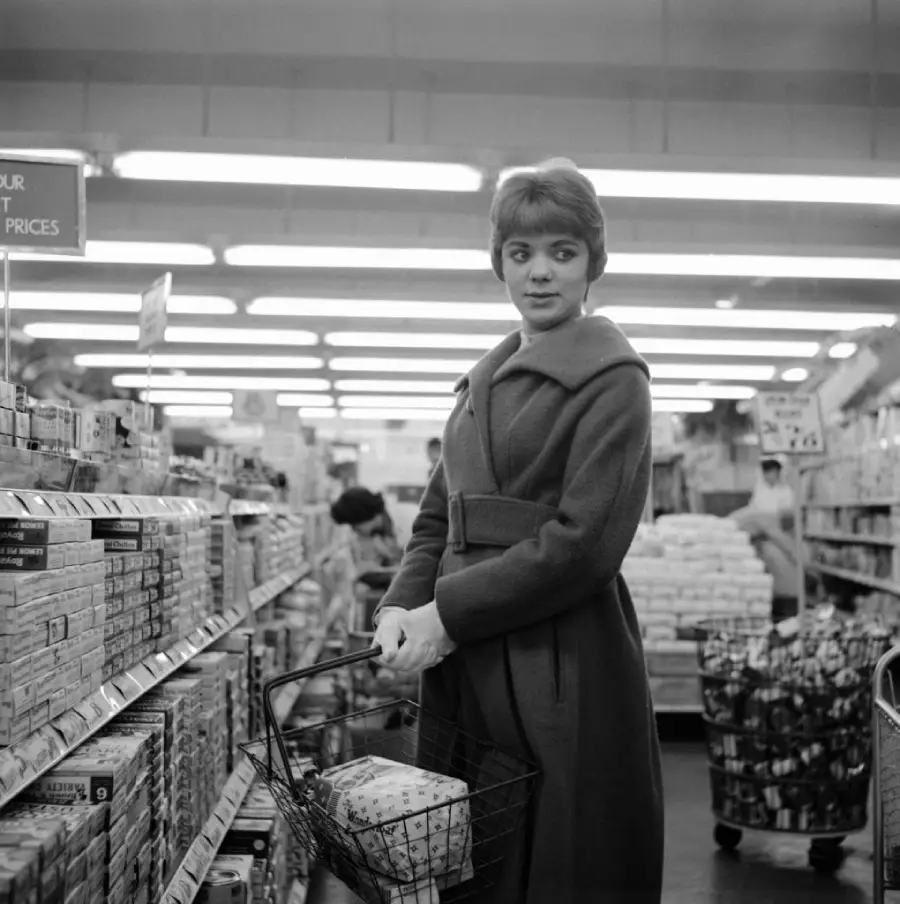 A woman in a coat holds a shopping basket in a grocery store aisle, looking over her shoulder. Shelves are stocked with various products, and other shoppers are visible in the background.