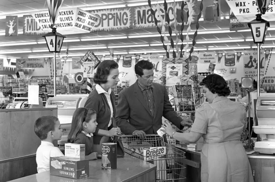 A family with two children stands at a supermarket checkout, interacting with a cashier. The scene appears vintage, with 1950s attire and décor, and grocery items in their cart and on the counter.