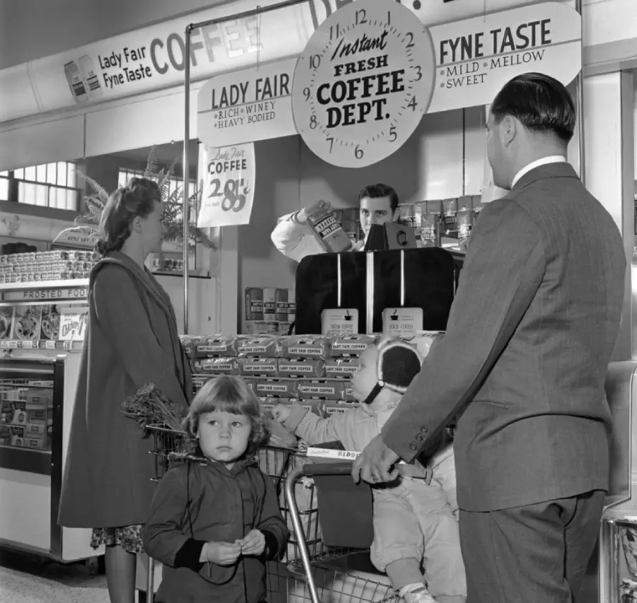 A family stands near a coffee display in a grocery store. A man behind the counter serves coffee, while a young girl and a woman stand beside a shopping cart with a small child inside. Signs advertise coffee prices and brands.