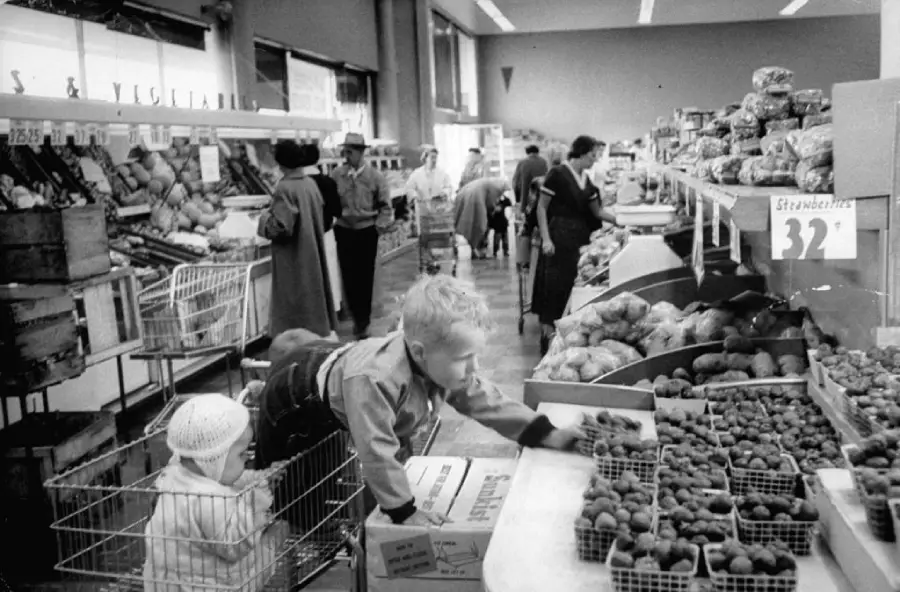 Black and white photo of a busy grocery store. A boy reaches for strawberries while a smaller child sits in a shopping cart. Shoppers in coats browse produce aisles in the background.