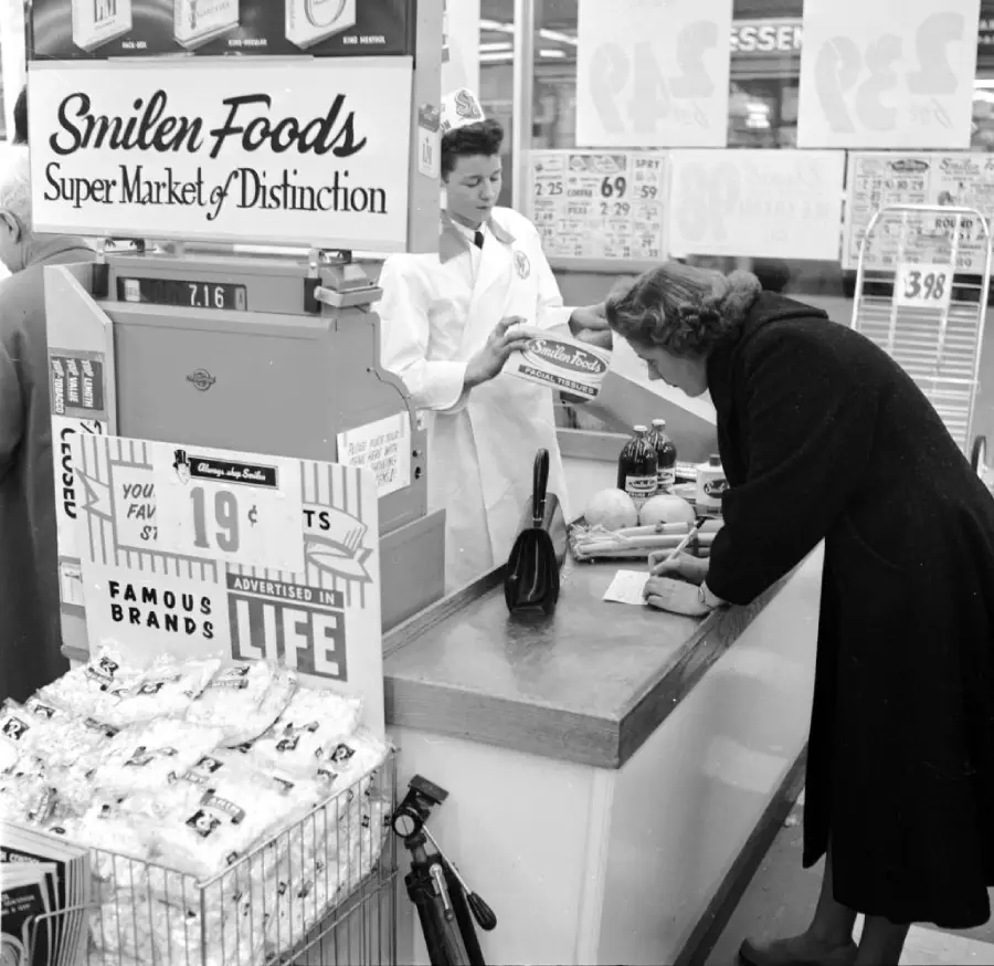 A woman at a supermarket checkout counter interacts with a cashier in uniform. Groceries, including produce and packaged goods, are on the counter. Signs and food packs are visible in the background.