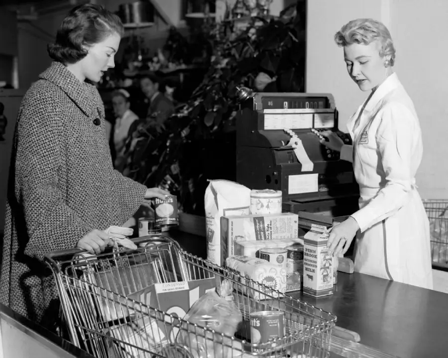 A woman in a coat stands at a grocery store checkout with a full cart, while a cashier in a uniform rings up items on a vintage cash register. The scene appears to be from the mid-20th century.