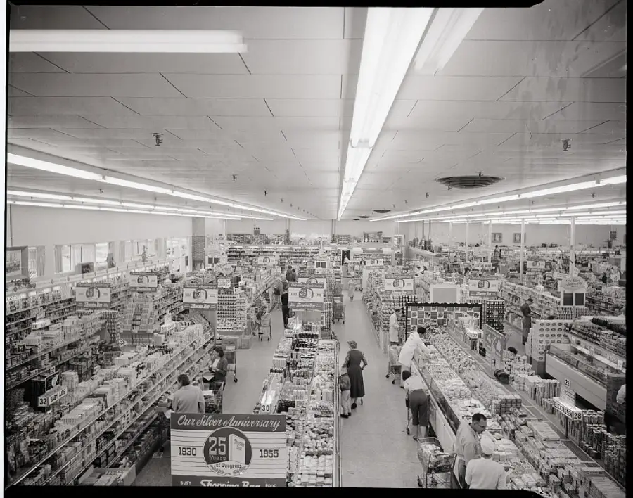 Black and white photo of a crowded supermarket with shoppers and employees among aisles of groceries. Anniversary sale signs are visible at the front of the image.