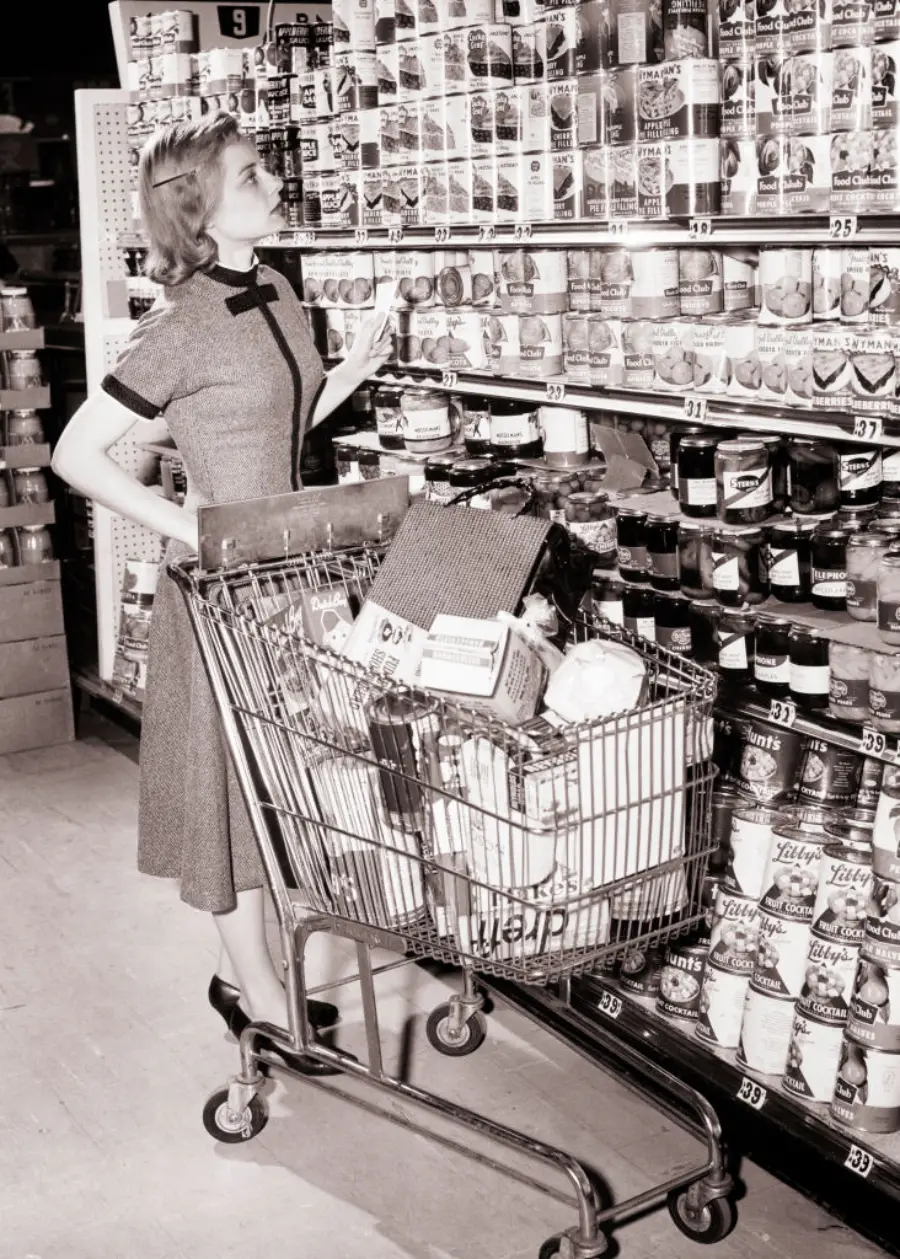 A woman in a vintage dress stands by a grocery cart full of items, reaching for canned goods on a supermarket shelf. The image is black and white, evoking a mid-20th-century setting.