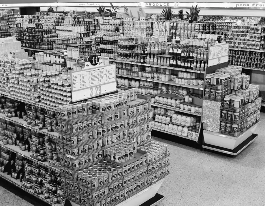 Black and white photo of a grocery store interior with shelves stocked with canned goods, jars, and various products, arranged in neat rows and displays on a tiled floor.