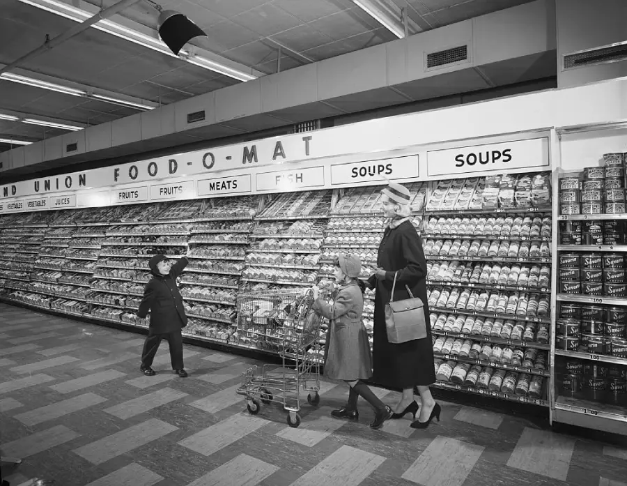 A woman and two children shop in a supermarket aisle filled with canned goods. The woman pushes a shopping cart while the children walk beside her. The shelves are neatly stocked and labeled with foods like meats, fruits, and soups.