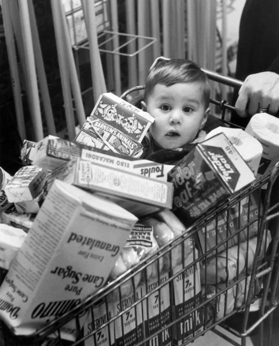 A young child sits inside a grocery cart filled with various food items, including cereal boxes and cartons. The child looks up with a surprised expression. An adult hand is seen pushing the cart.