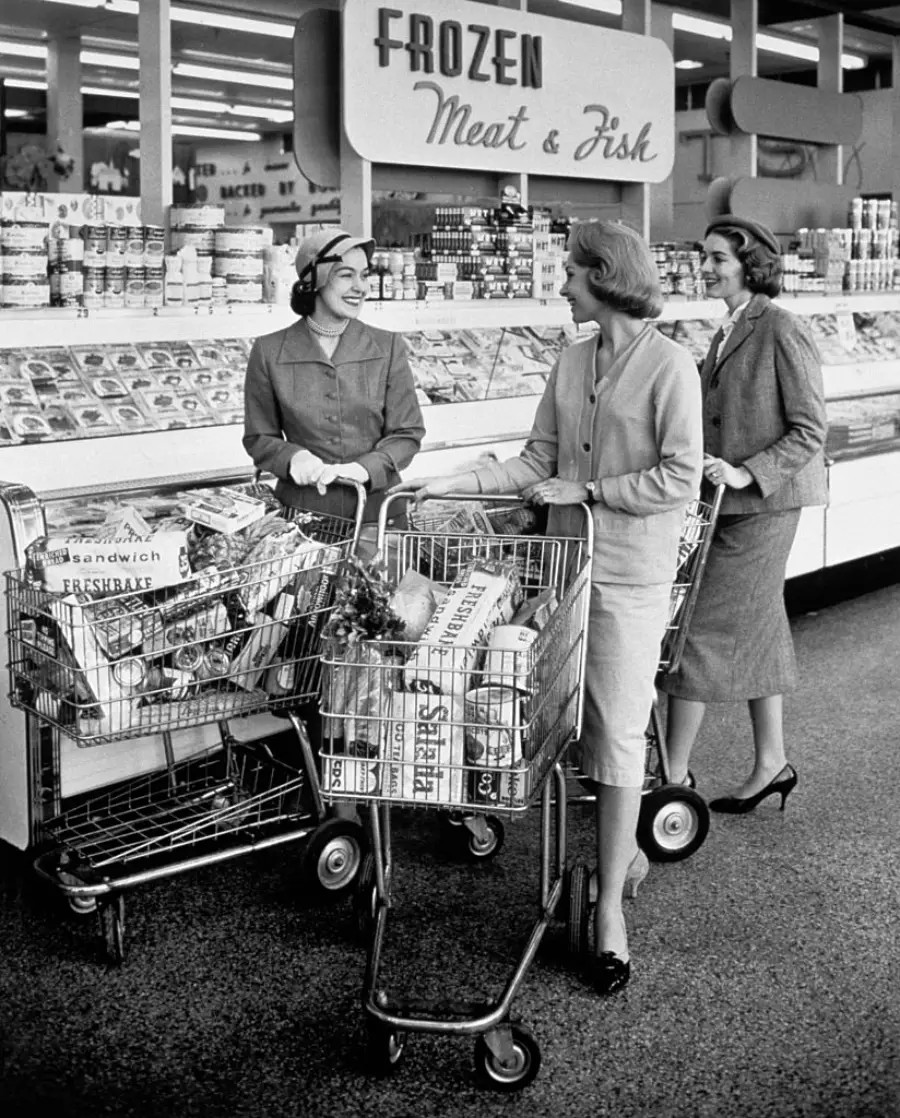 Three women in 1950s-style clothing chat and smile while shopping in a grocery store aisle, each with a full cart. A sign behind them reads “FROZEN Meat & Fish.”