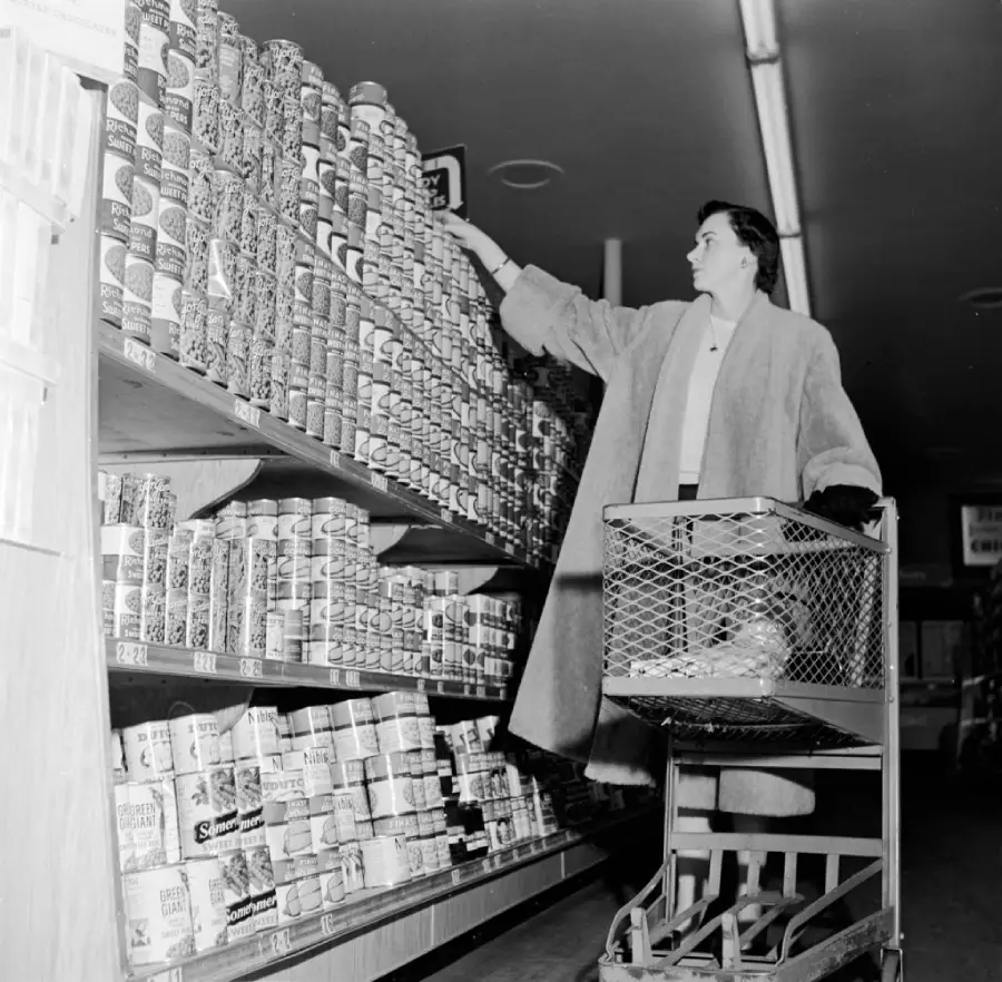 A woman wearing a long coat stands beside a grocery cart, reaching up to grab a can from a tall shelf stocked with canned goods in a grocery store aisle.