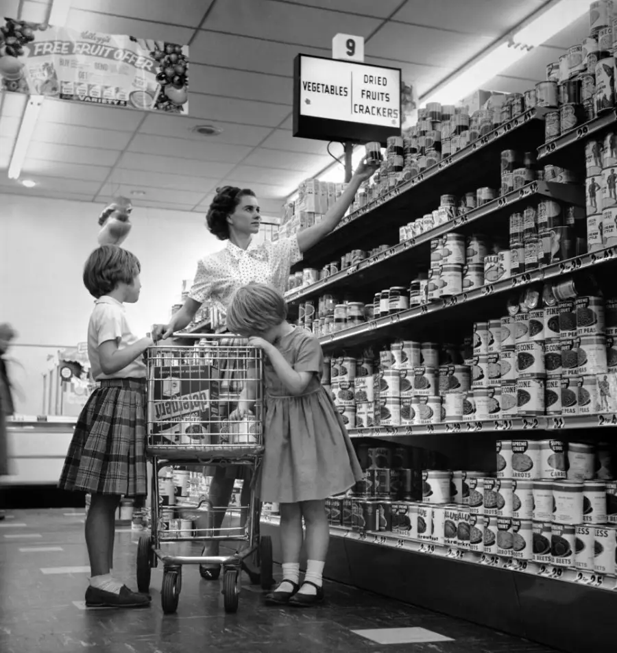A woman shops with two young girls in a grocery store aisle, reaching for a can on a shelf. The girls stand by a shopping cart. The aisle sign above reads “Vegetables, Dried Fruits, Crackers.”