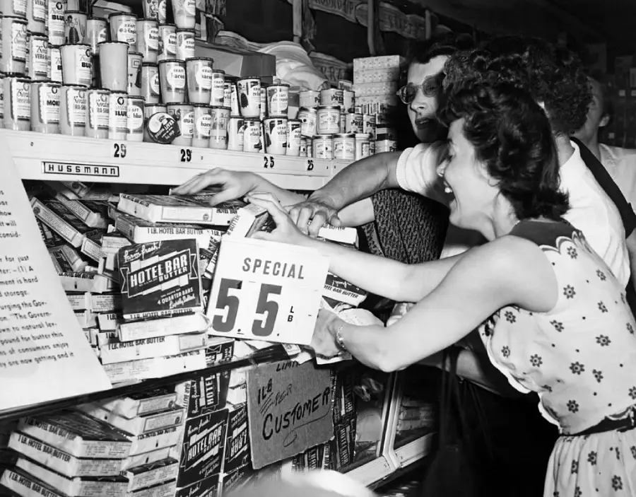 Black and white photo of shoppers eagerly grabbing butter from a grocery store display marked “SPECIAL 5.5.” Canned goods are stacked above, and signs with prices are visible on the shelves.
