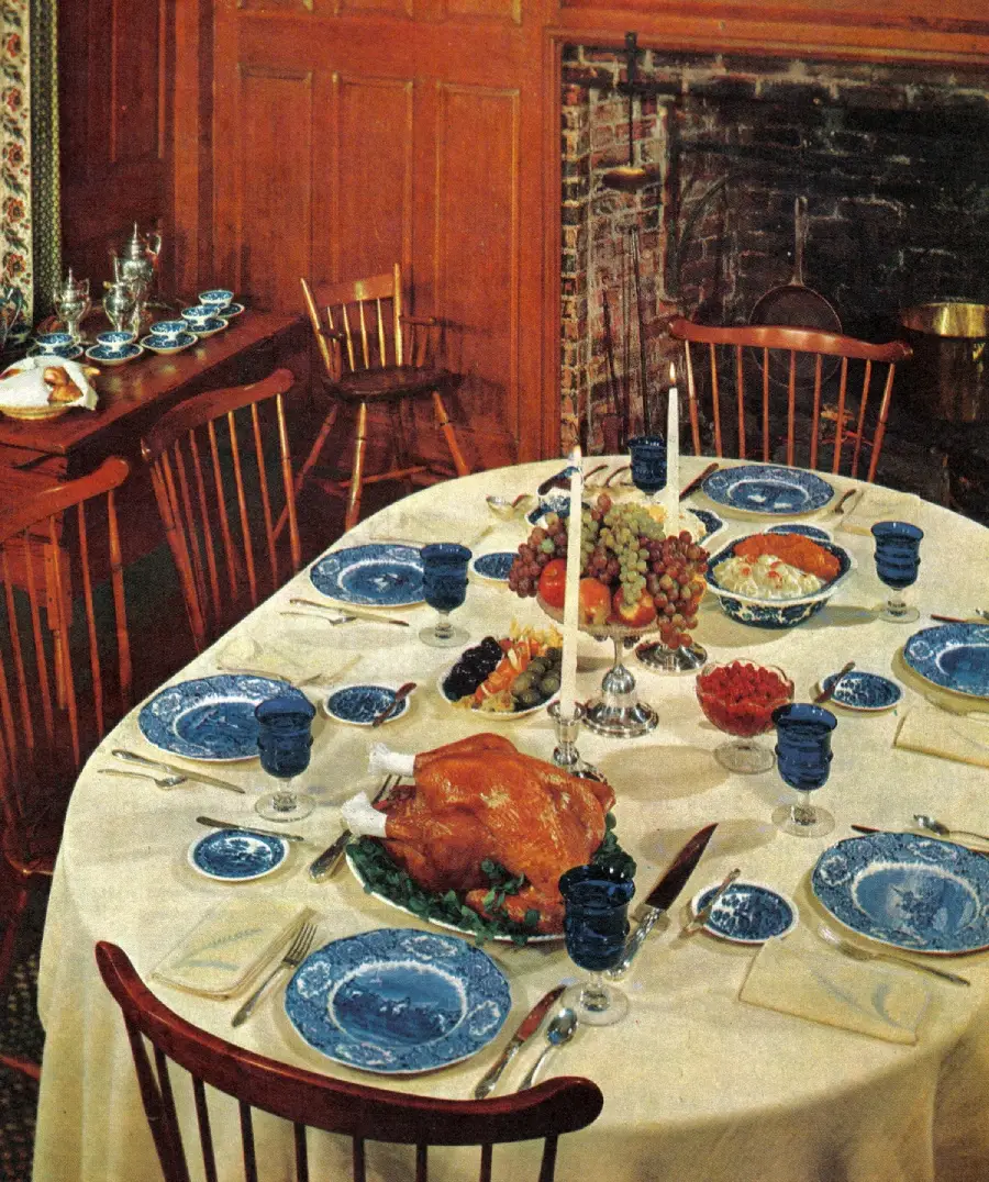 A dining table set for a meal with blue dishes, glasses, a roast turkey, fruit, and candles, surrounded by wooden chairs in a cozy room with a fireplace and wood paneling.
