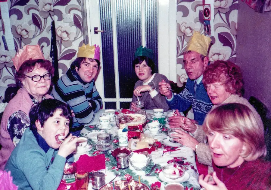 Seven people of various ages sit around a table, eating and wearing colorful paper crowns, likely celebrating a holiday, in a room with floral wallpaper and a festive tablecloth.