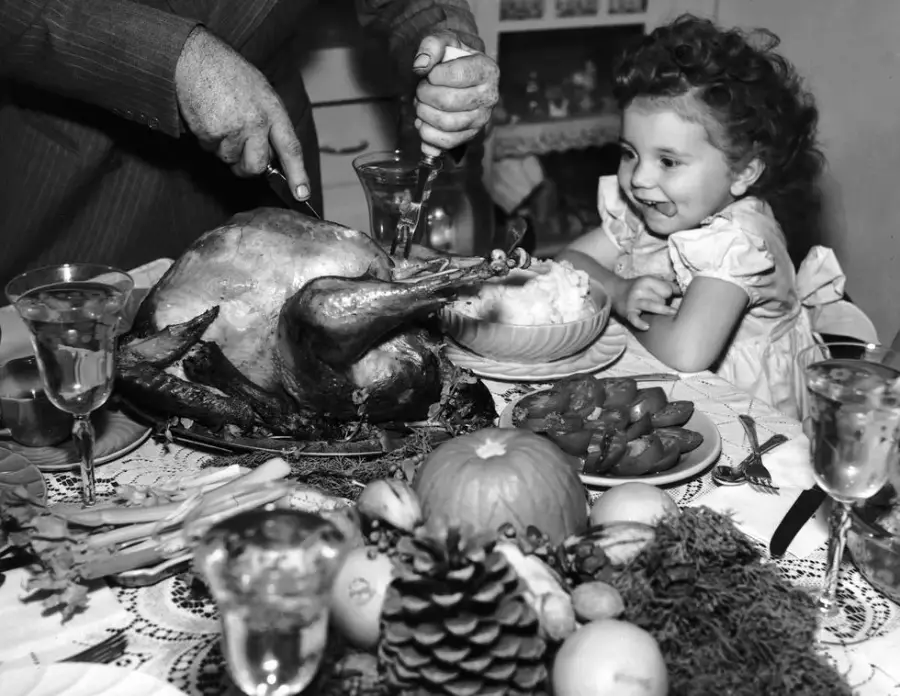 A smiling child sits at a table as an adult carves a large roasted turkey. The table is set with vegetables, fruit, and glasses, creating a festive, cozy atmosphere.