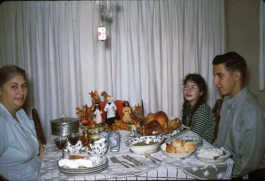 Three people sit at a dining table set with a roast turkey, rolls, and various dishes. Festive autumn decorations are in the center, and the group appears ready for a meal, likely Thanksgiving.