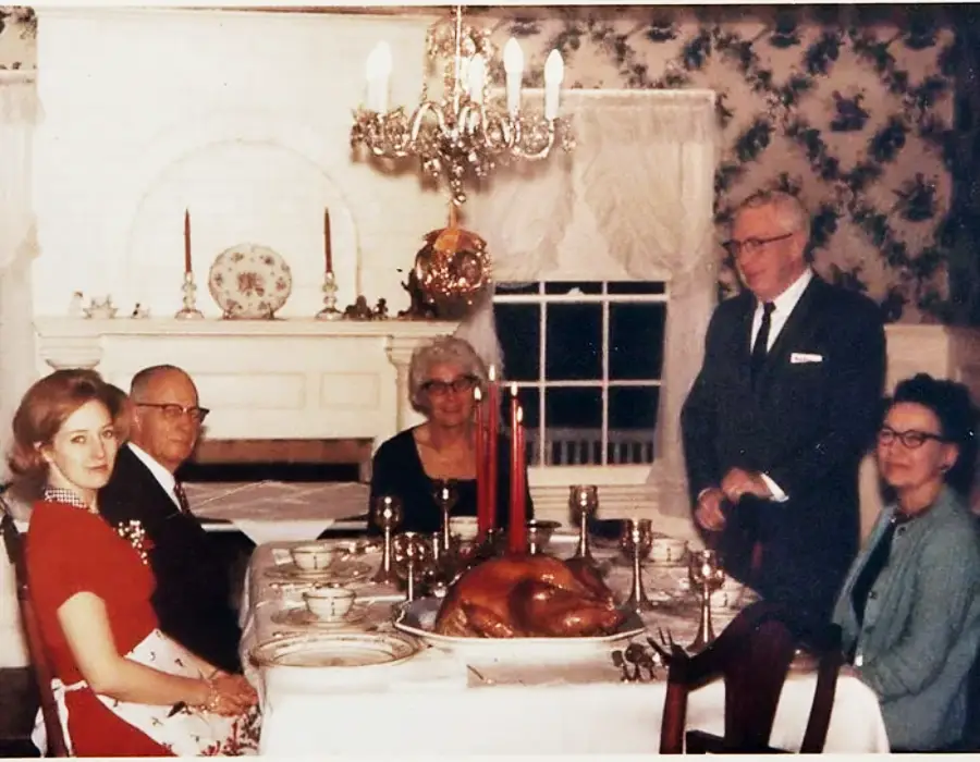 Five adults sit or stand around a dining table with a roast turkey centerpiece in a vintage-style dining room. The group is dressed formally and appears to be gathering for a special meal.