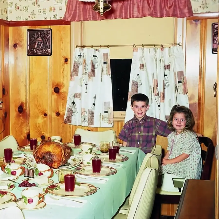 Two children sit at a dining table set for a meal with a roast turkey, drinks, and dishes. The room has wood-paneled walls, patterned curtains, and vintage decor, giving a cozy, retro atmosphere.